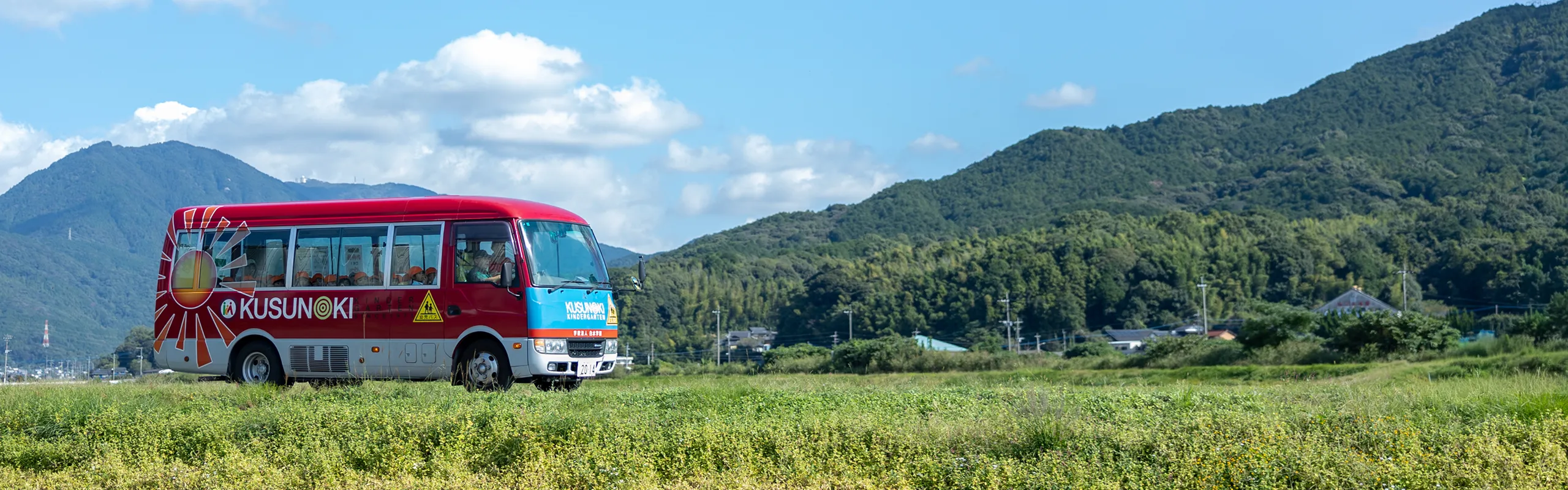 山々を背景に田園を走るくすの木幼稚園の送迎バス